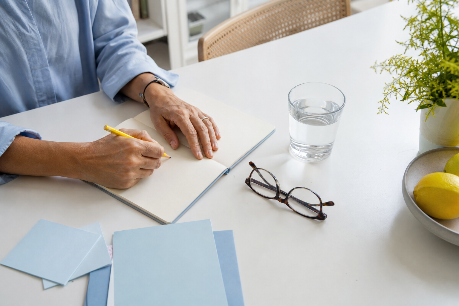 Persoon schrijft vragen in een notitieboek naast een glas water en een bril